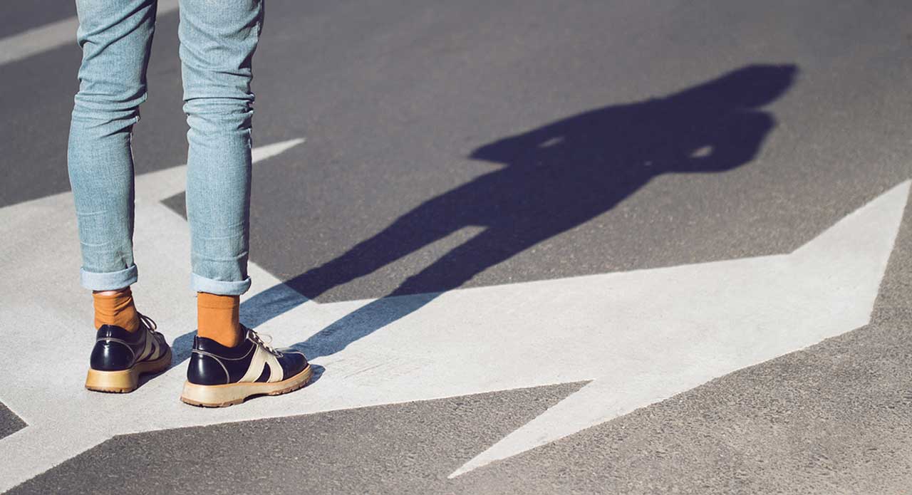 Photo of a person's shadow at the edge of a road, with two arrows pointing in opposite directions